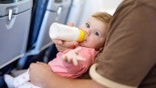 Baby girl seen on dad's lap on airplane. She is drinking from bottle and has her arm outstretched toward camera