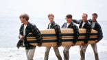 The Beach Boys smiling wearing matching plaid shirts and beige pants holding a surfboard and walking on the beach.