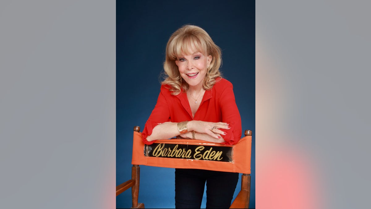 Barbara Eden in a red shirt and black pants posing in front of a chair with her name.