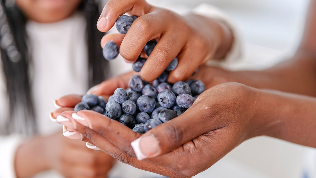 A Black woman holds blueberries in her hand while a young Black girl grabs a clump with her hand.