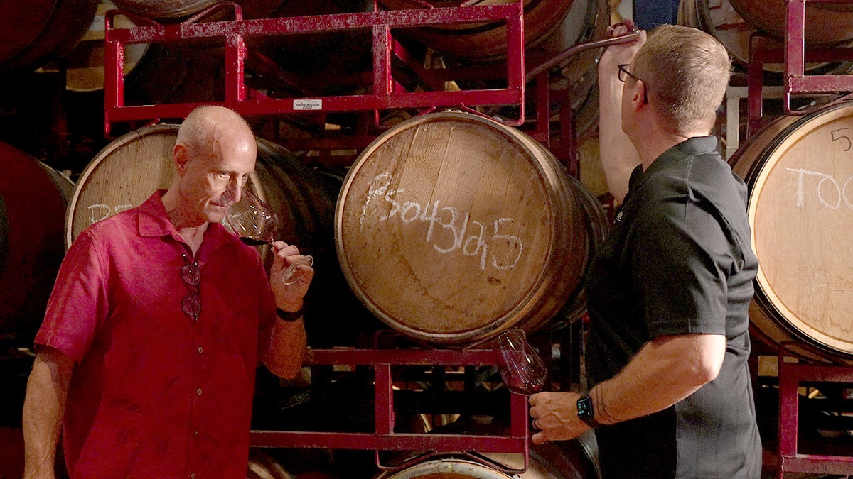 A winemaker raises a glass to his nose while an employee at a winery checks on a barrel.