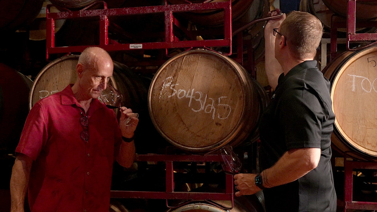 A winemaker raises a glass to his nose while an employee at a winery checks on a barrel.