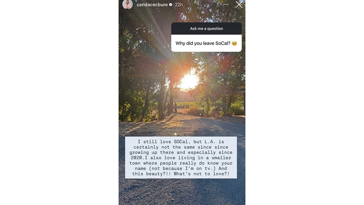 A sunset shines through trees onto a gravel path where two people stand, with lush greenery and fields in the background.