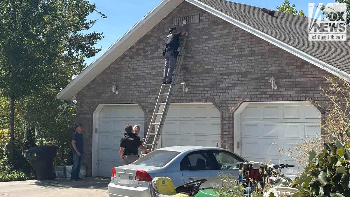 A uniformed police officer on top of a ladder removing a white security camera from above a garage door