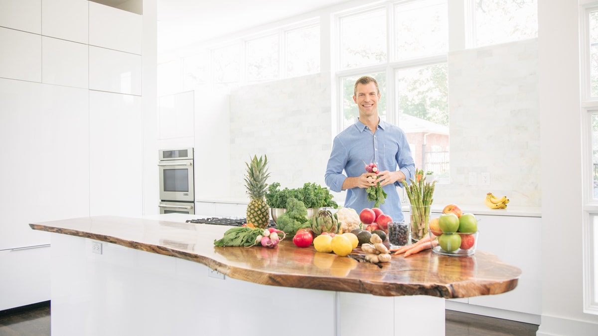 Dr. Ax stands in a white kitchen with a table covered with fruits and vegetables