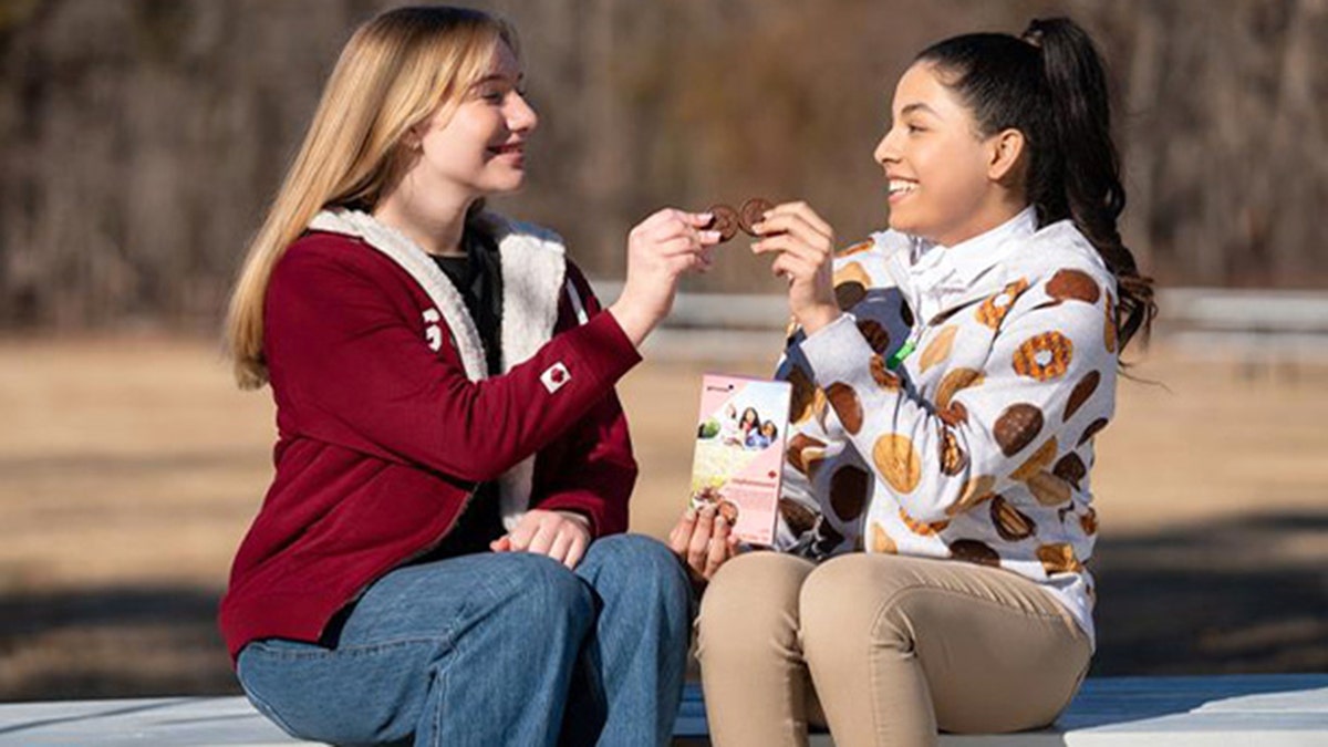 Two older girls are sitting outside and holding Exploremores, one of the new Girl Scout cookies, while the one one on the right holds the box.