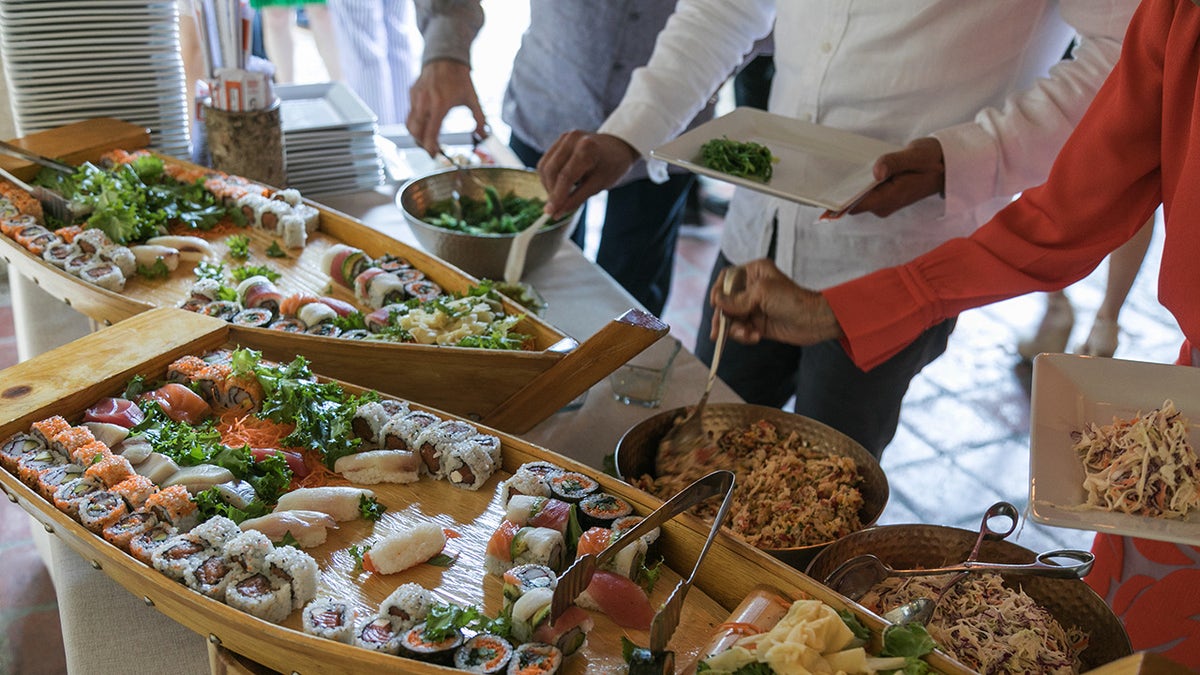 Three people reaching toward sushi bar at buffet, seen filling their plates with boats of sushi and sashimi in front of them