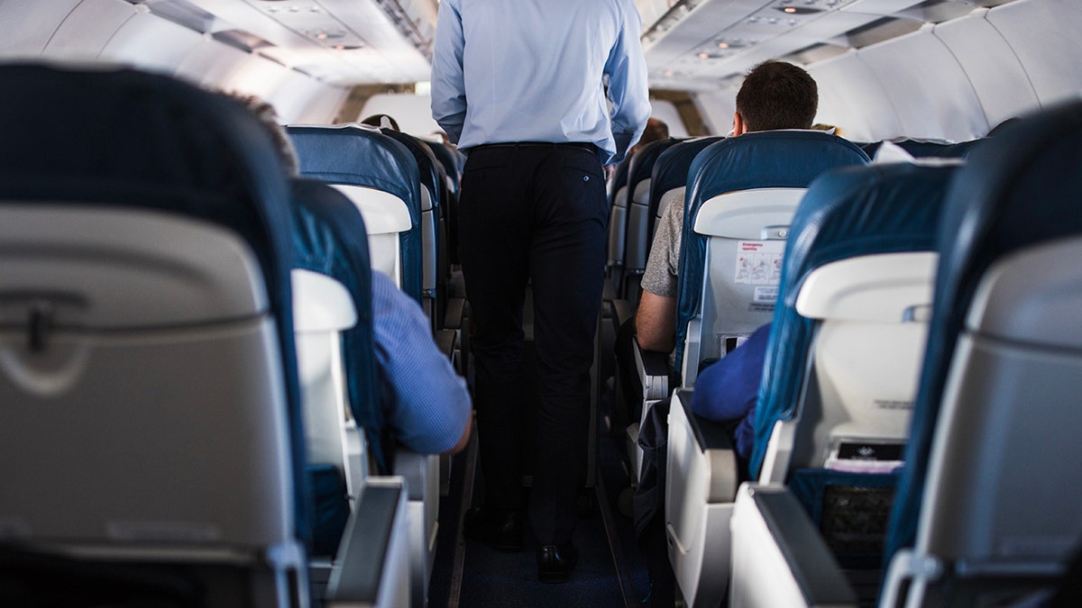 Flight attendant seen from behind walking down aisle with passengers in seats on both sides.