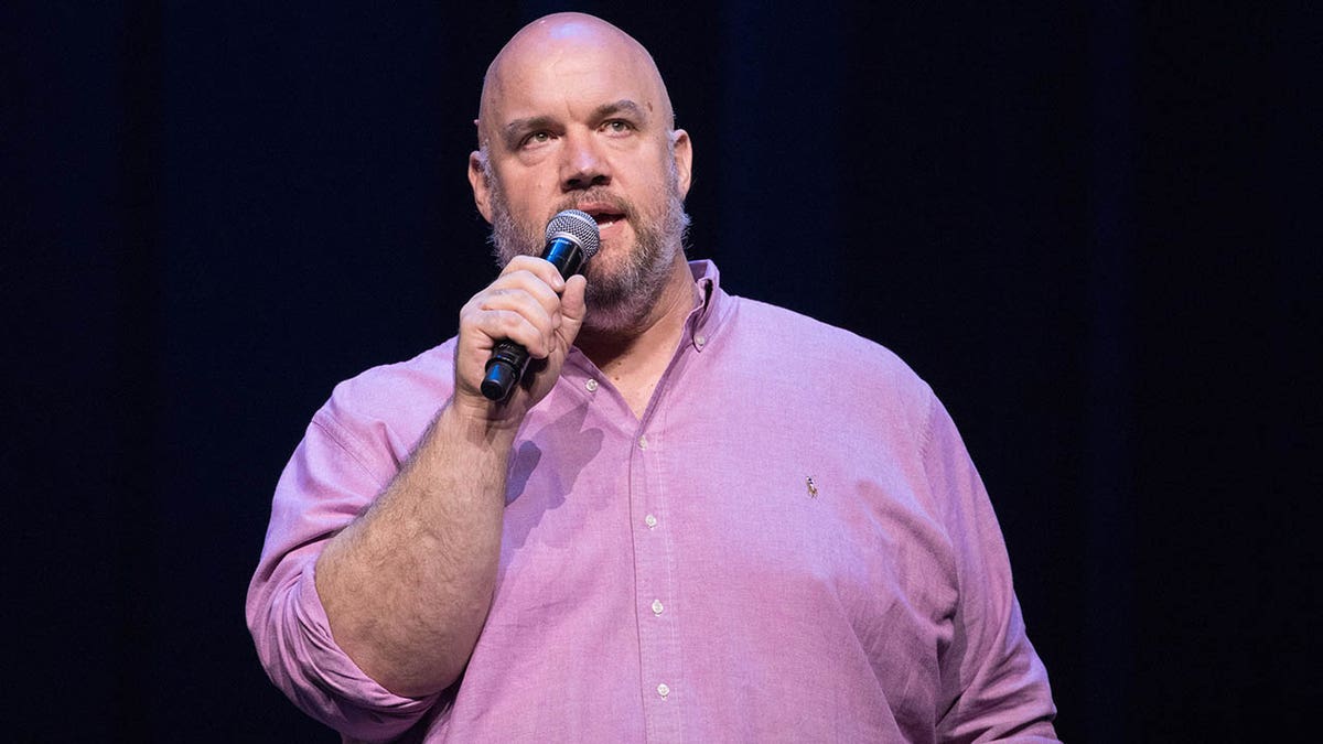 Guy Branum holds a microphone on stage.