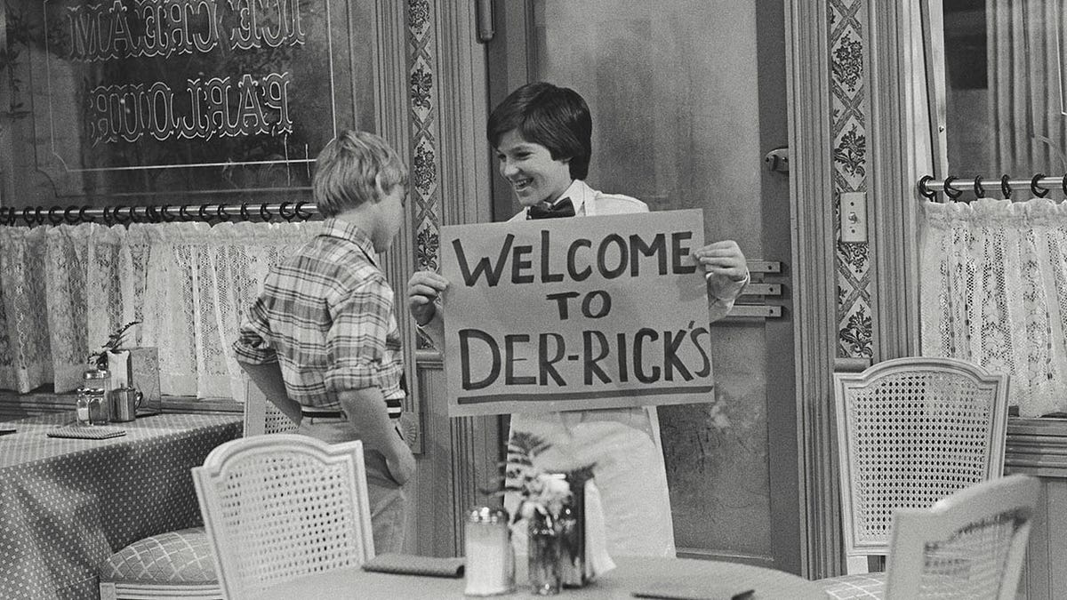 Jason Bateman, Silver Spoons'tan bir sahnede bir işaret tutar.