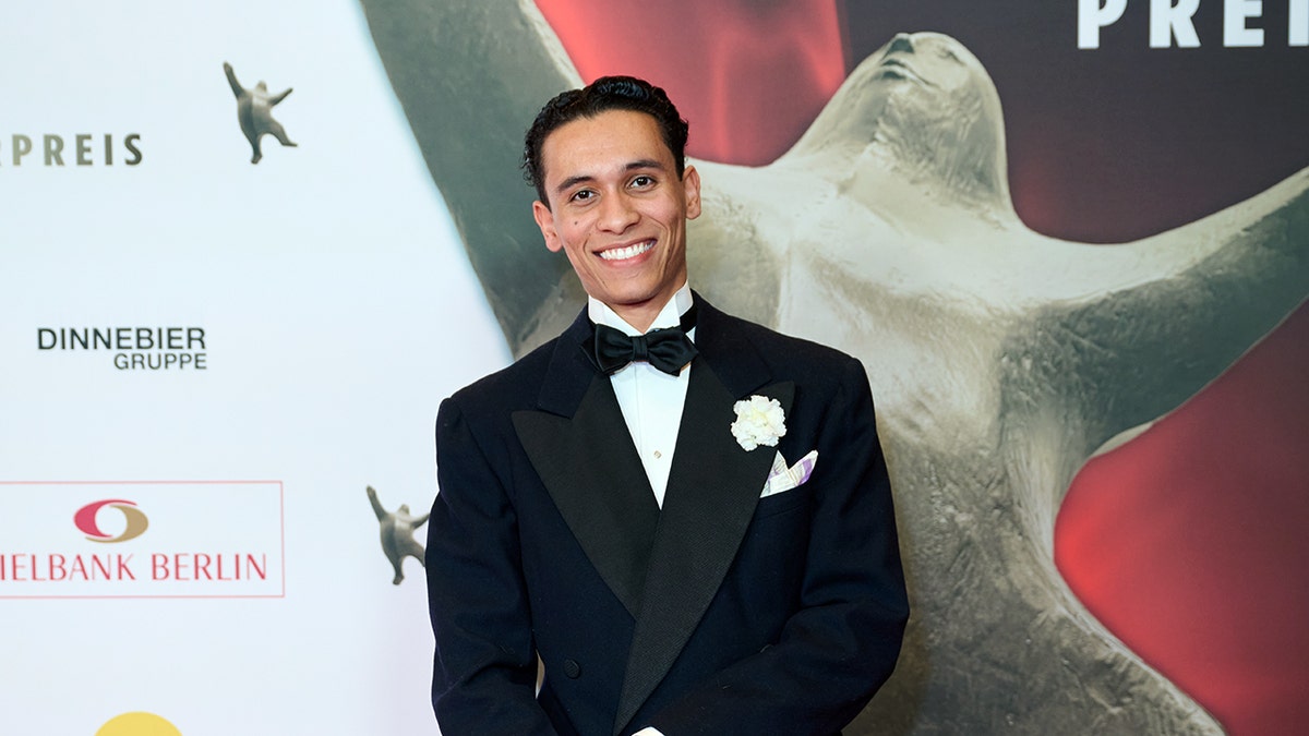 David Hermlin smiles on red carpet in tuxedo with bow tie and flower lapel.