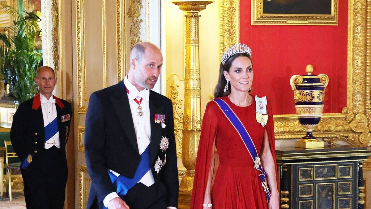 Prince William and Kate Middleton in formal royal wear walking inside the palace in front of Prince Edward.