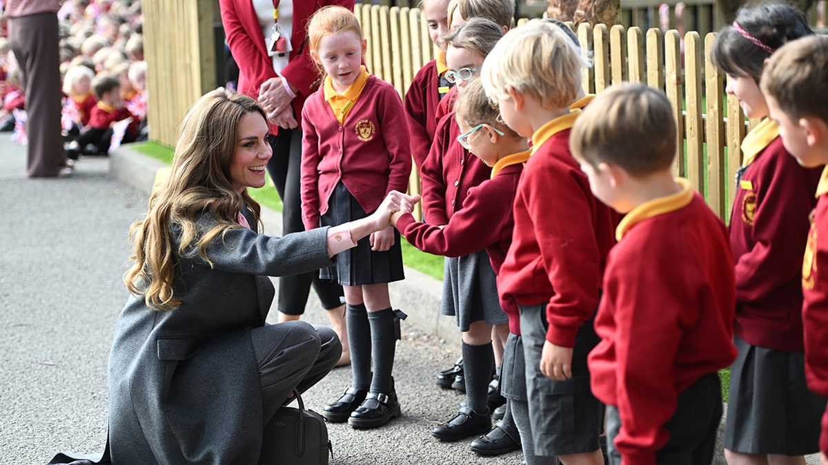 Kate Middleton on her knees greeting children while smiling at them.