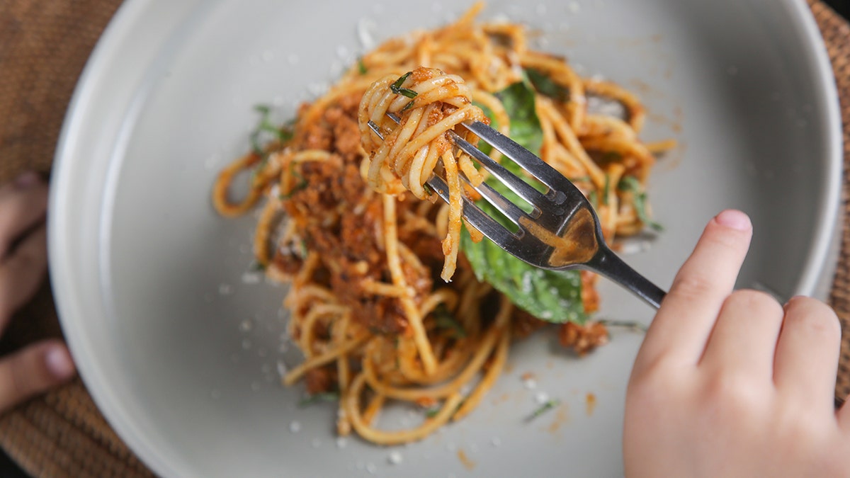 Little girl eating plate of spaghetti with tomato and meat sauce, seen from above with just her hands as she twirls pasta with fork