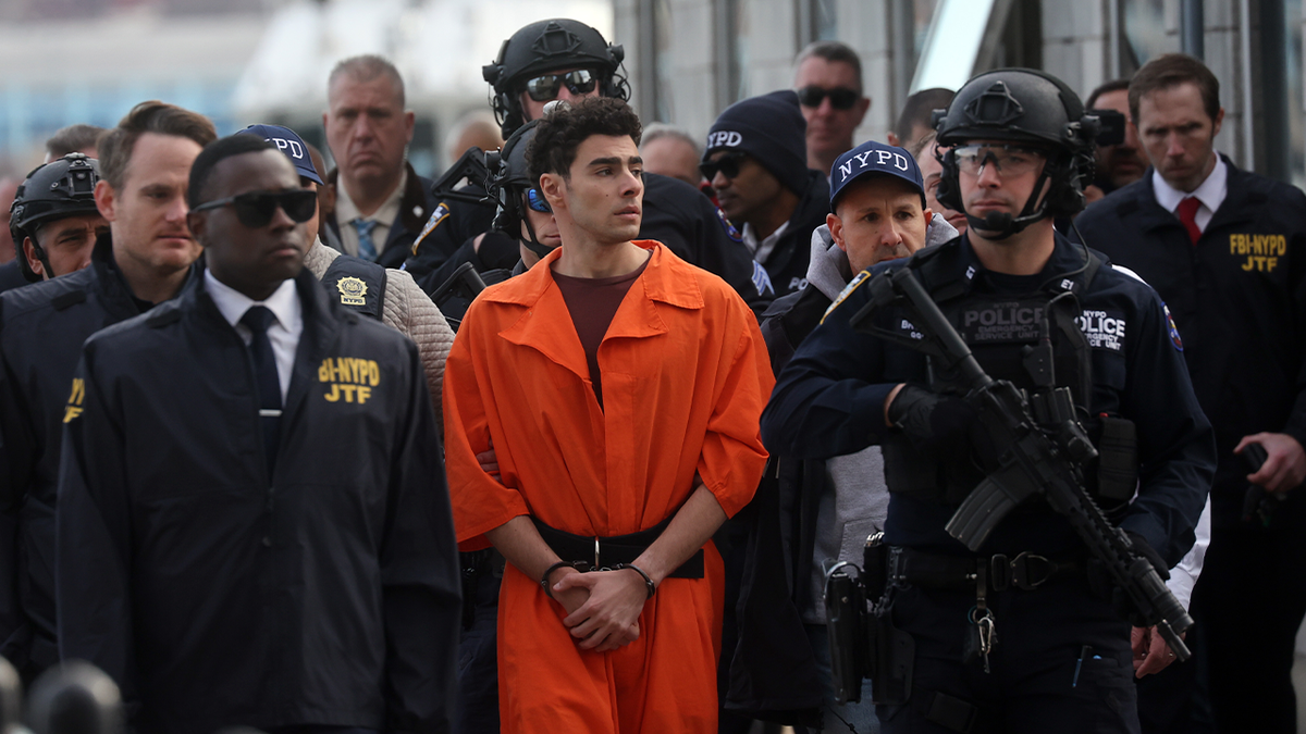Luigi Mangione in an orange inmate jumpsuit flanked by armed members of the NYPD as they escort him away from the helicopter that carried him part of the way back to NYC following his extradition from Pennsylvania