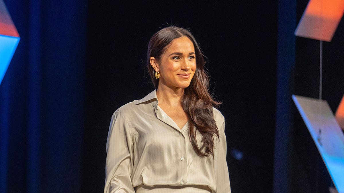 Meghan Markle wearing beige walking on stage smiling.