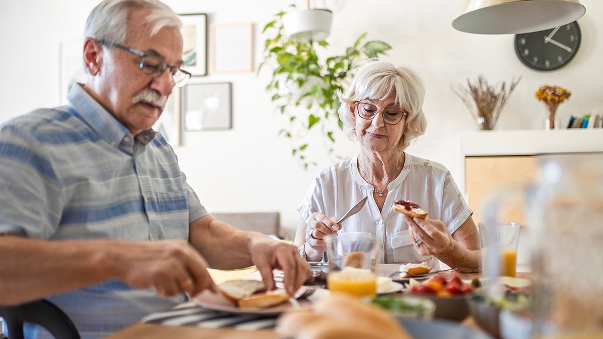 O casal sênior toma café da manhã em casa, coloca geléia na mesa da cozinha com outros alimentos e suco de frutas na frente deles, mostrando a dieta saudável e equilibrada necessária para aumentar a saúde do coração.