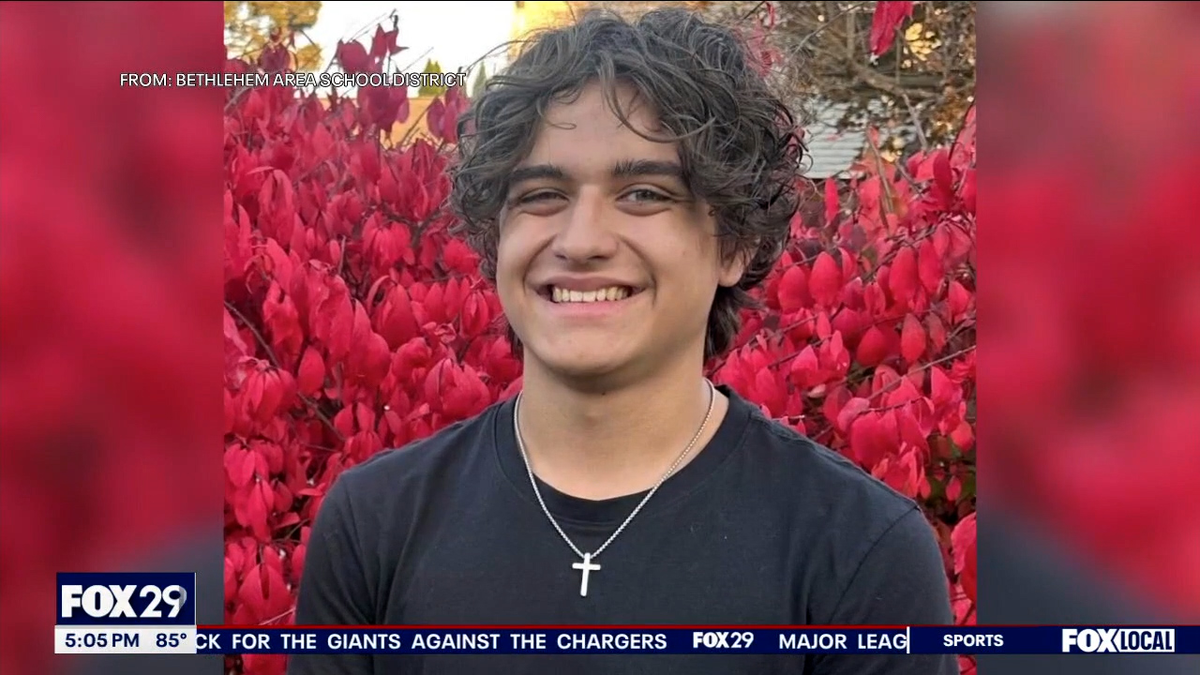 An image showing David Nagy, wearing a black T-shirt with a cross necklaces, standing in front of a large bush full of red flowers