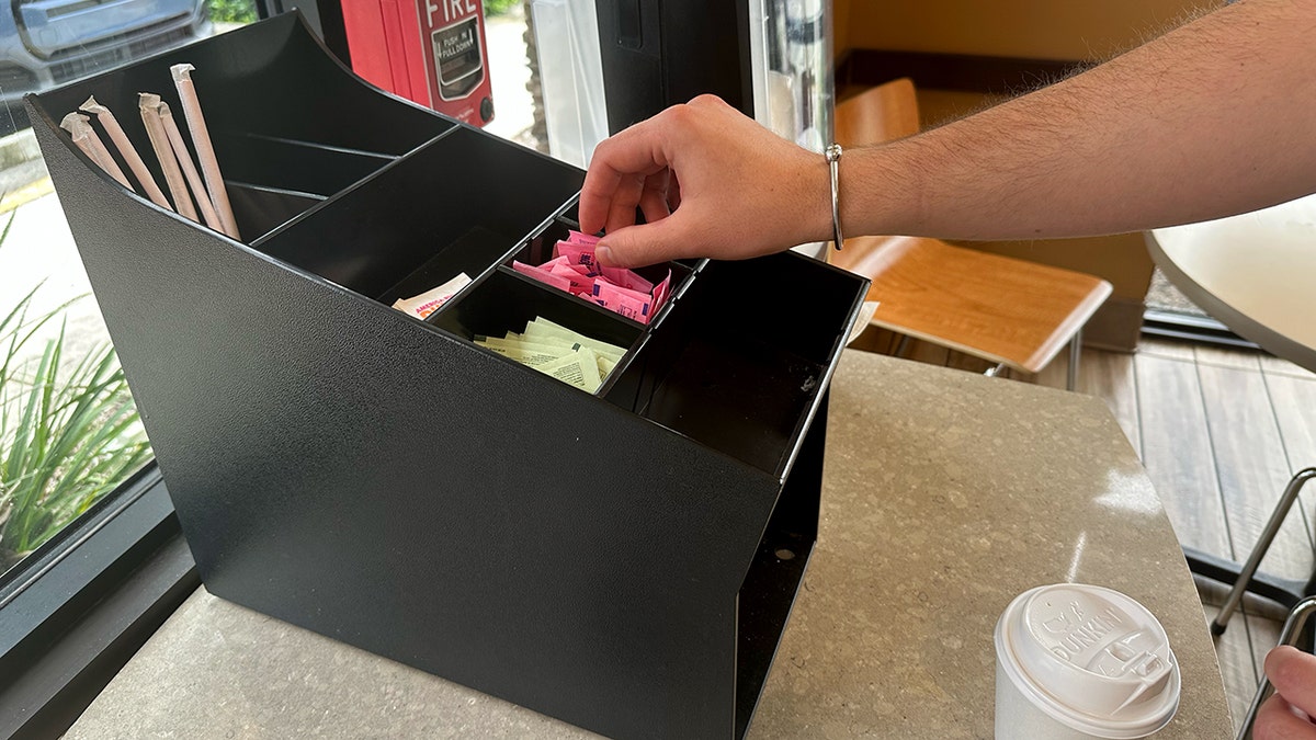 A hand reaches for packets of Sweet 'N Low at a Dunkin'.