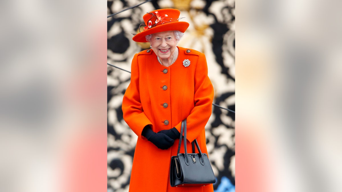 Queen Elizabeth wearing a bright orange coat dress as she holds a black purse.