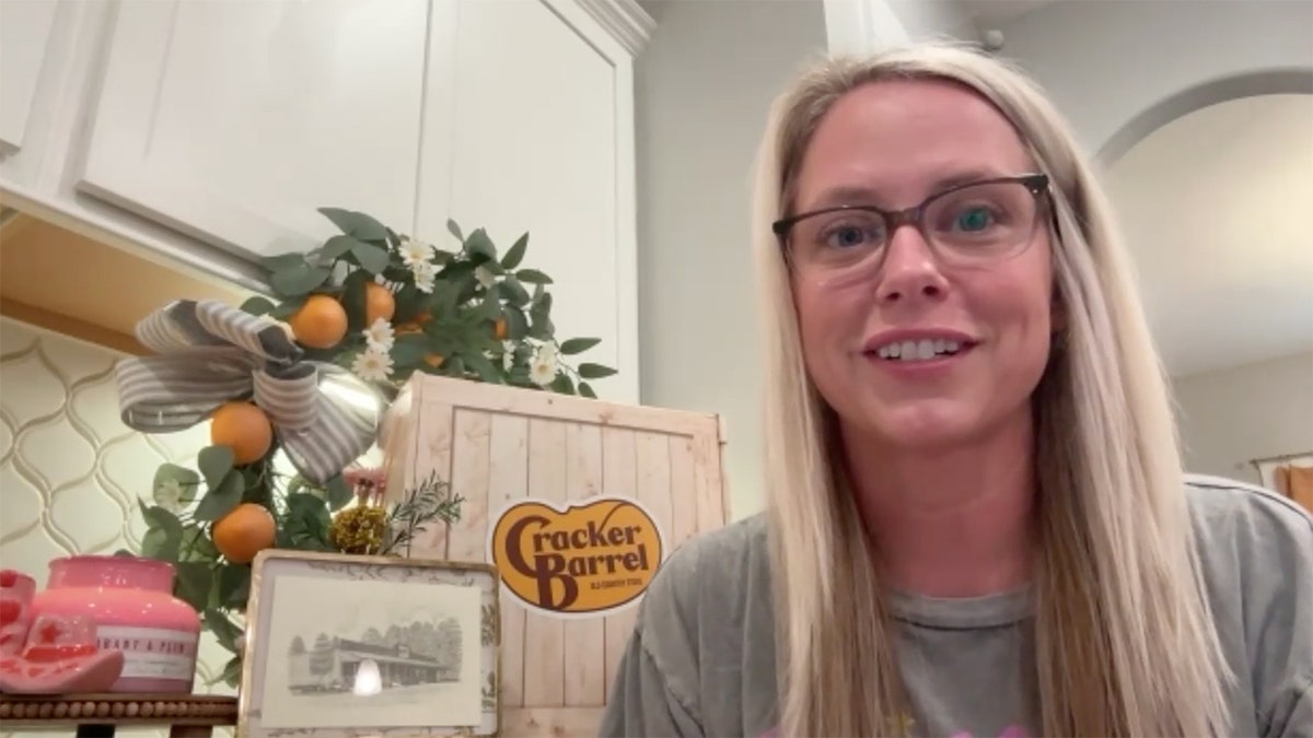 A blonde woman sits next to a Cracker Barrel gift box and a framed drawing of one of its stores.