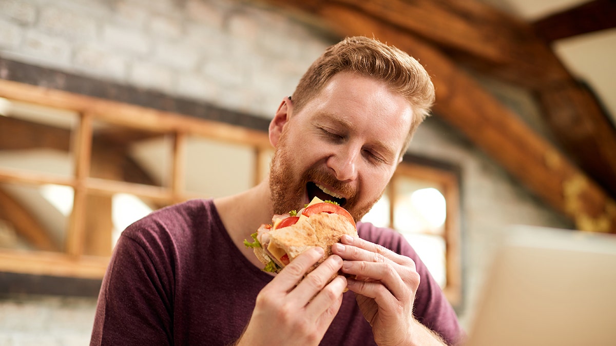 Man with eyes closed eating sandwich for breakfast at home.