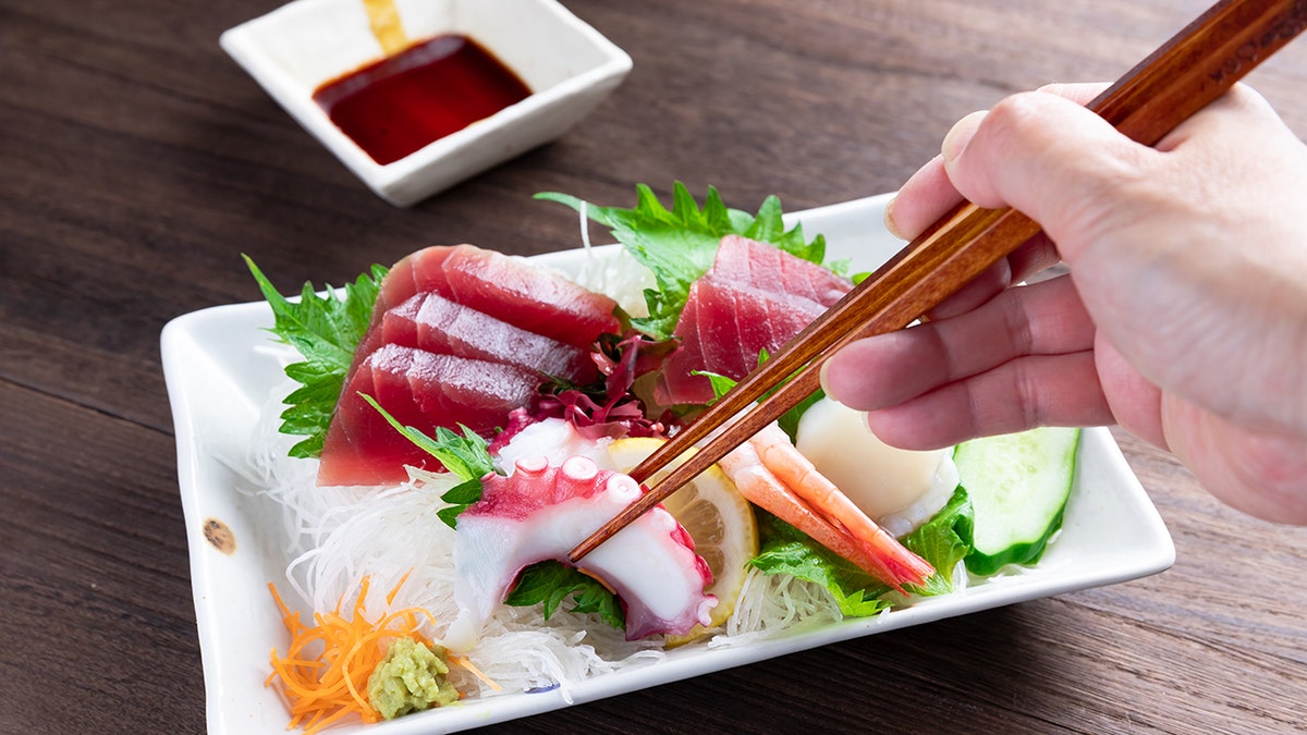 Hand with chopsticks reaching for piece of octopus on plate of Japanese sashimi with dipping cause on side.