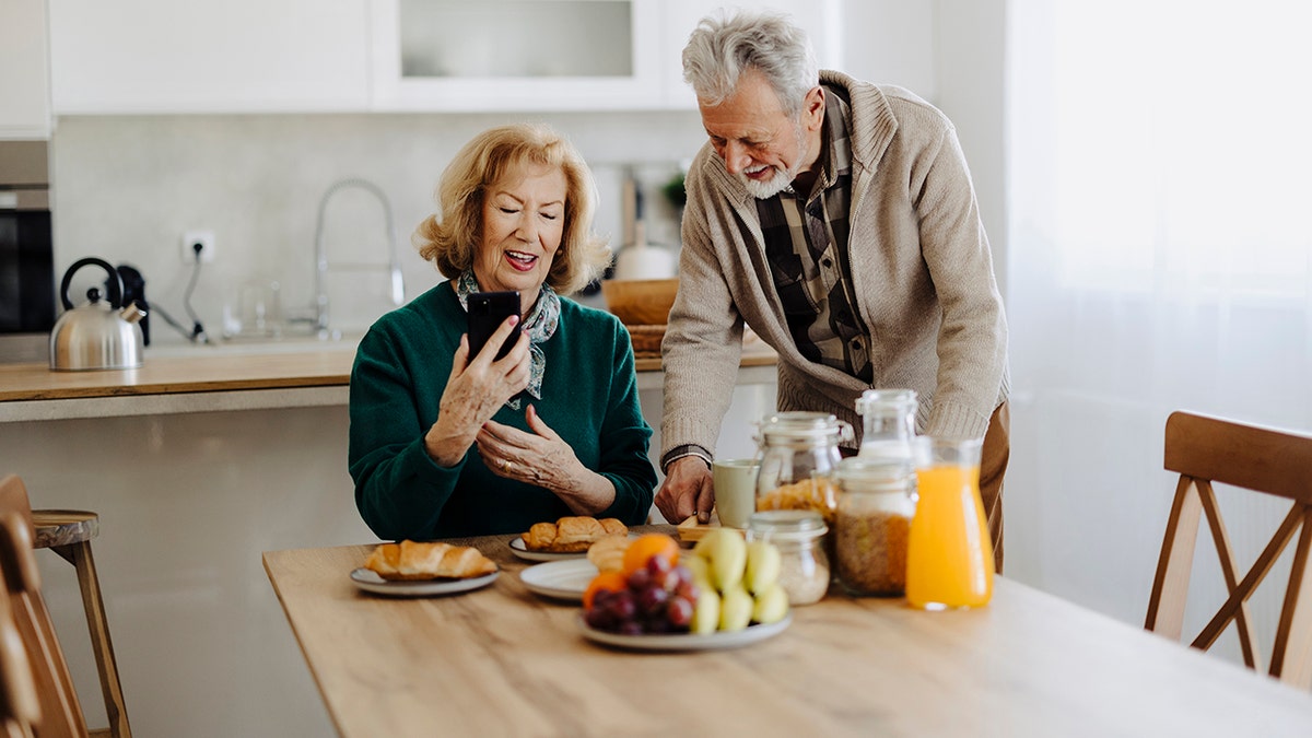 A happy couple having healthy breakfast together as his wife showed her what was on her phone and he stood beside her and smiled. The orange juice and fruit spread in breakfast.