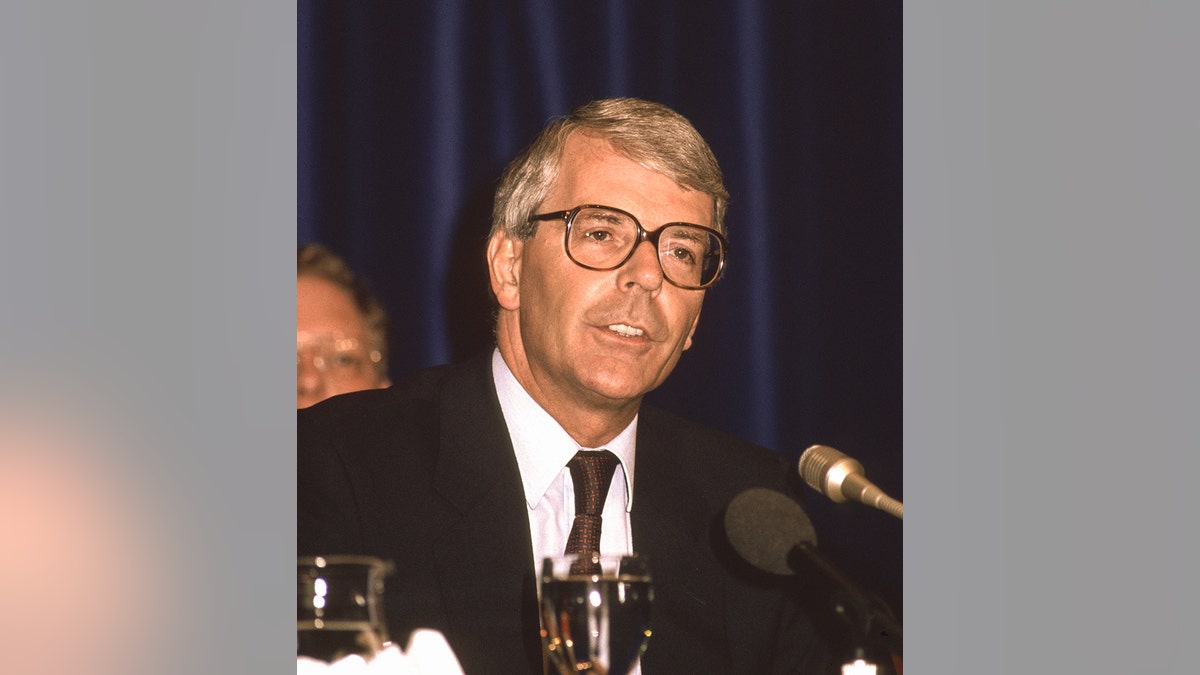 John Major in a suit speaking at a podium.