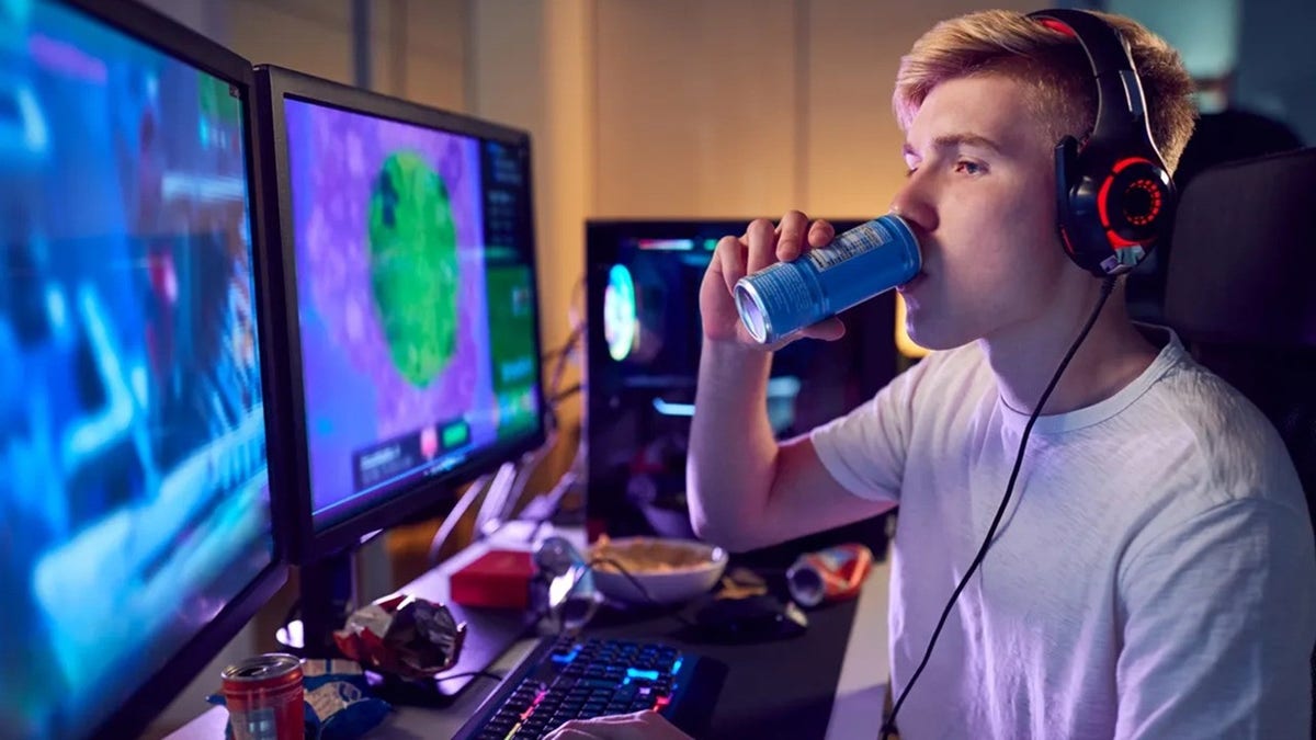 Teenaged boy playing video games on computer screens with headphones on, drinking energy drink