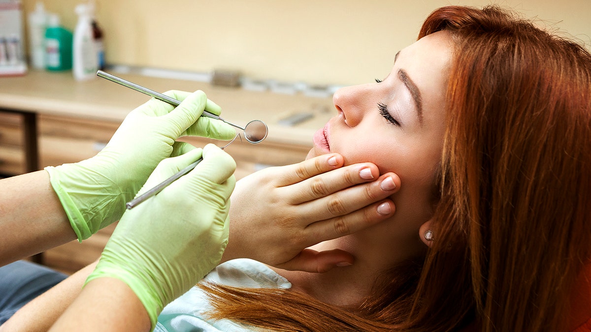 Teenaged girl at dentist, holding face indicating toothache as dentist reaches toward her with tools