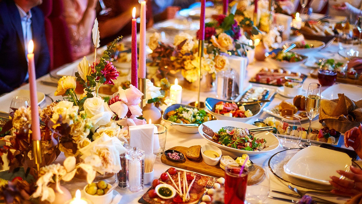 Wedding reception food spread out on decorated table with guests at edges. Different plates of meats, cheeses, vegetables and other appetizers seen along with drinks including glasses of champagne