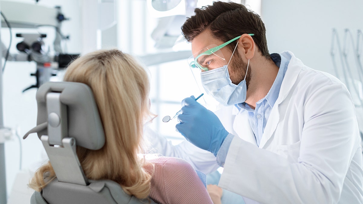 A woman gets her teeth examined by male dentist seen wearing mask and goggles and holding instruments