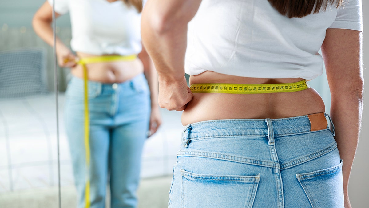 Young woman wearing jeans and white tee taking measurements of her body in front of the mirror in her bedroom, measuring tape seen around her stomach