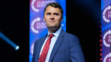 Charlie Kirk in a blue suit and red tie, clean shaved, on statge in front of a pair of Turning Point USA signs