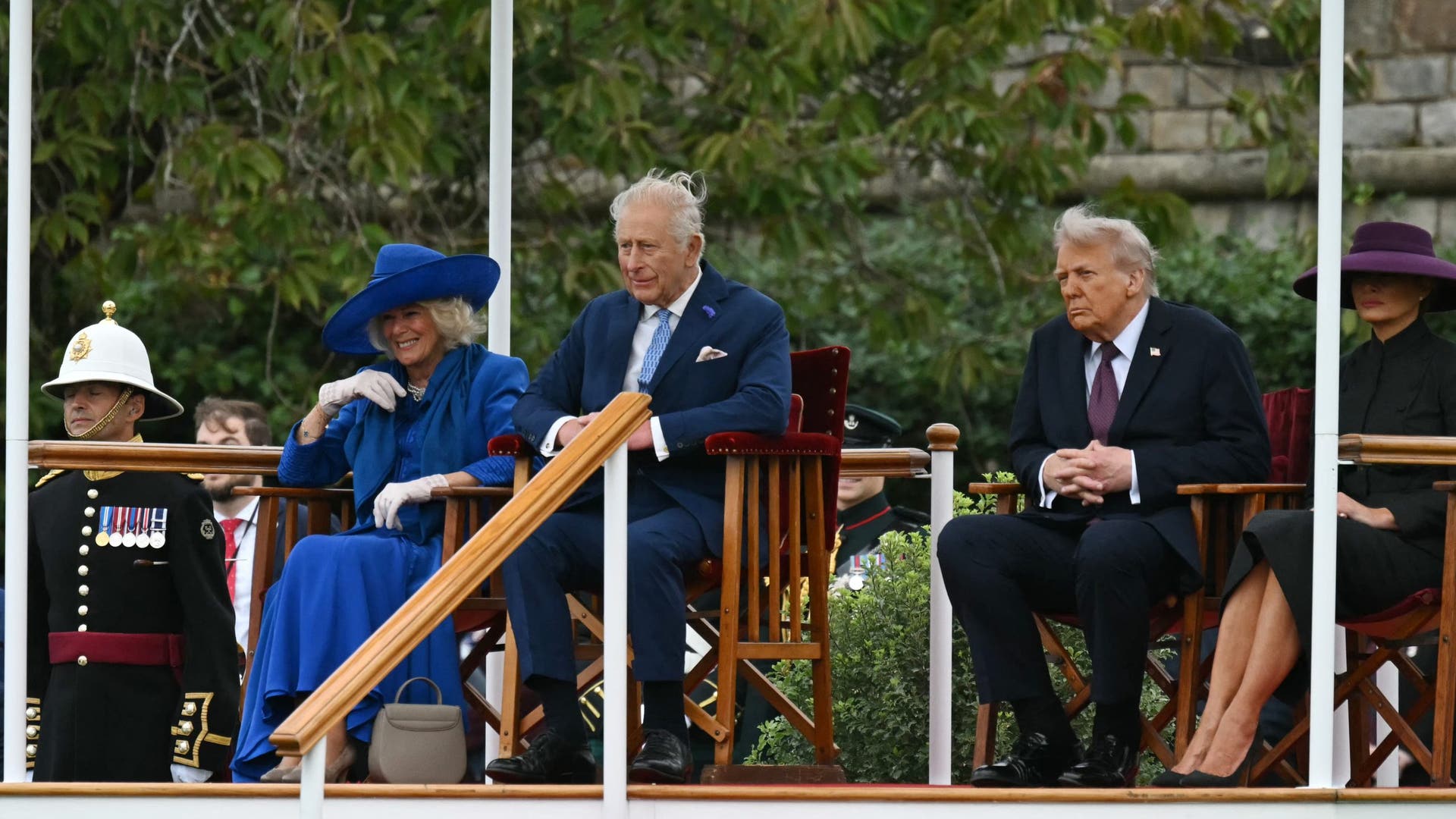 Queen Camilla smiles in a bright blue outfit as she sits next to King Charles III and Donald Trump during a royal ceremony.