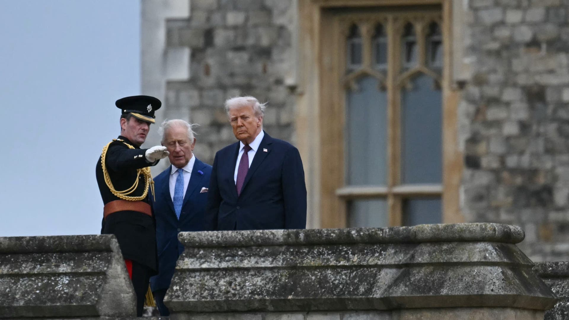 King Charles and Donald Trump listen to a British officer in full dress uniform pointing across the castle grounds.