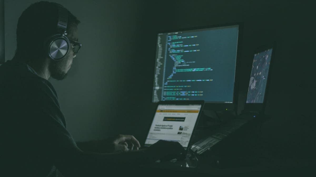 A person wearing headphones sits in a dark room, working on multiple computer screens displaying code.
