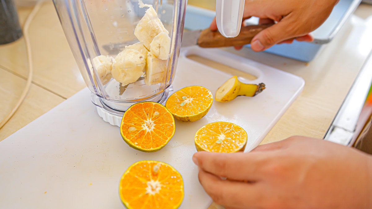 Persons hands seen cutting oranges to add to a smoothie, with banana already in blender.