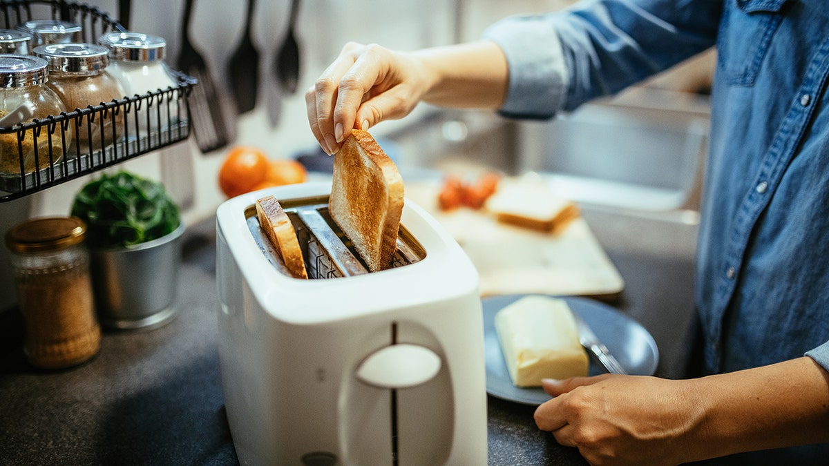 Woman's hands seen pulling toasted bread from toaster, butter and other incredients on counter around her.