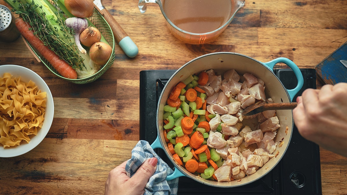 Person stirring chicken and vegetables in a pot to make soup. Broth, veggies and noodles laid out on counter around the cook.