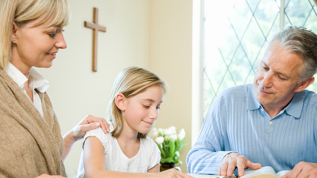 Foto stock de uma família multigeracional lendo a Bíblia à mesa com uma cruz na parede ao fundo.