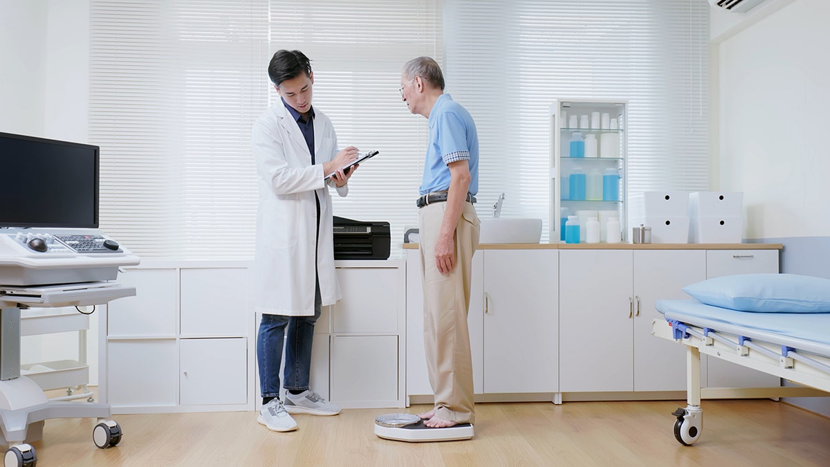 Doctor analyzing an older man’s weight as he stands on a scale during a medical examination.