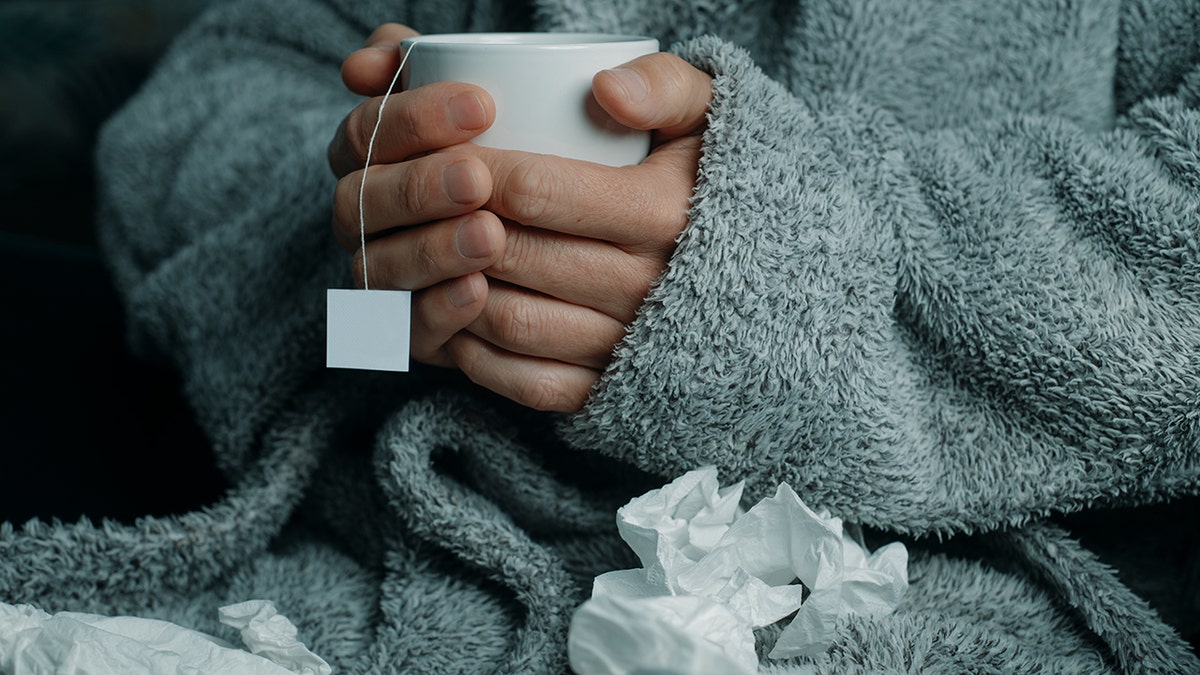 an ill man at home, wearing a fluffy gray house robe, surrounded by used tissues, warms up with a cup of hot herbal tea
