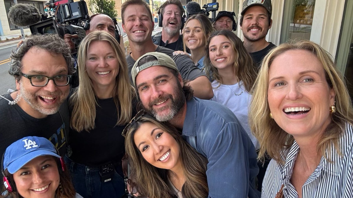 Group selfie of "Duck Dynasty" cast smiling with film cameras and crew equipment visible in the background.