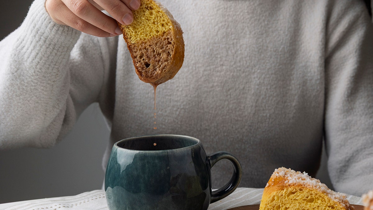 Woman dunking a piece of pumpkin bread into hot chocolate, seen up close as it drips into mug.
