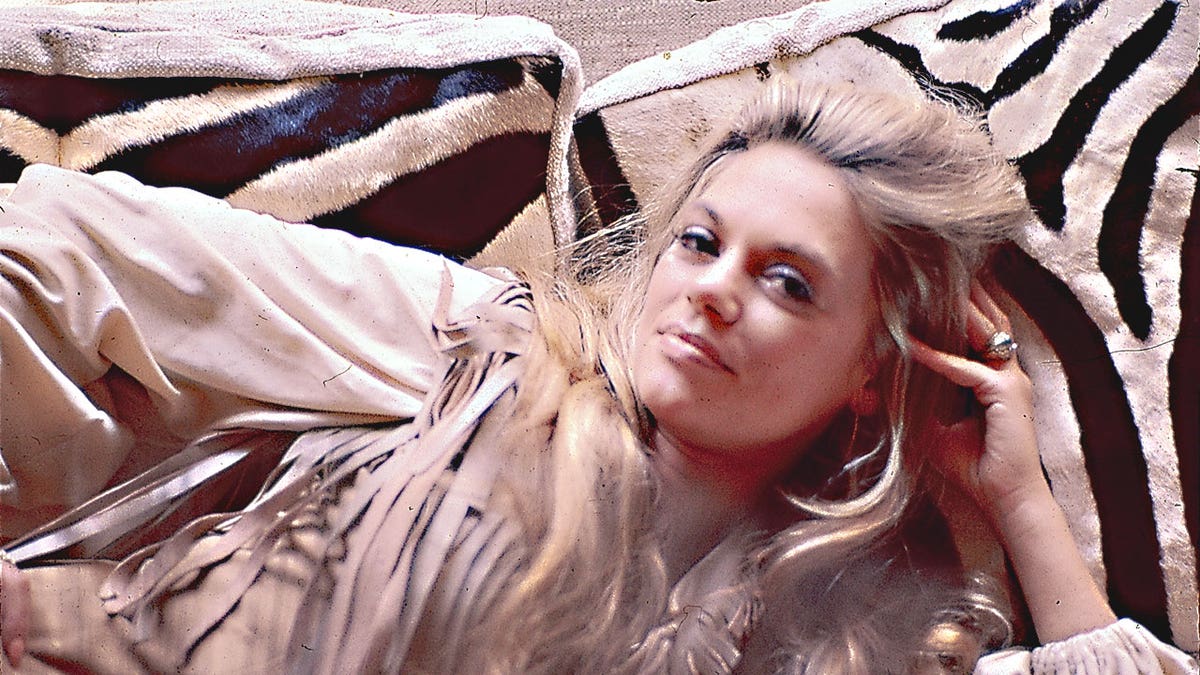Dyan Cannon wearing a beige flowy dress laying on a zebra bed.