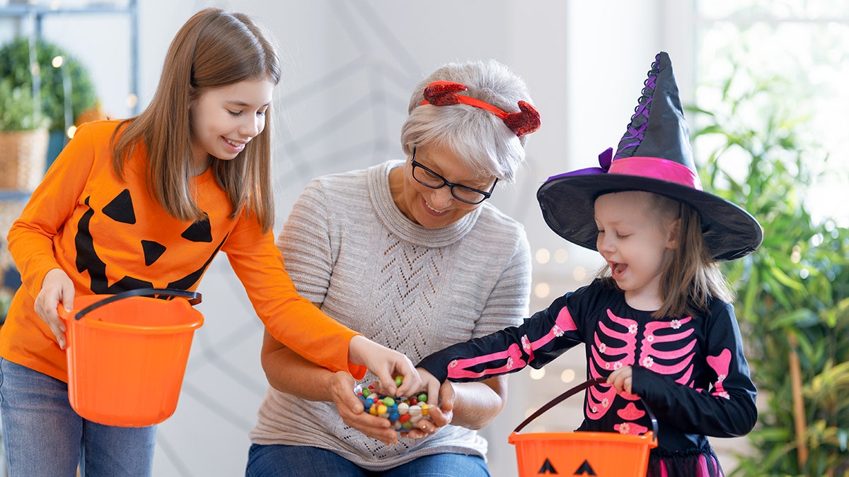 一位老婦微笑著拿著一碗糖果,裡面有兩個穿著萬聖節服裝的女孩手牽著手。