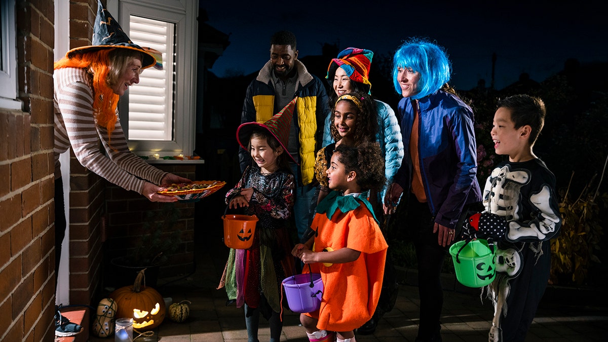 Older woman in witch hat handing out candy from doorway to trick-or-treaters dressed in colorful costumes.