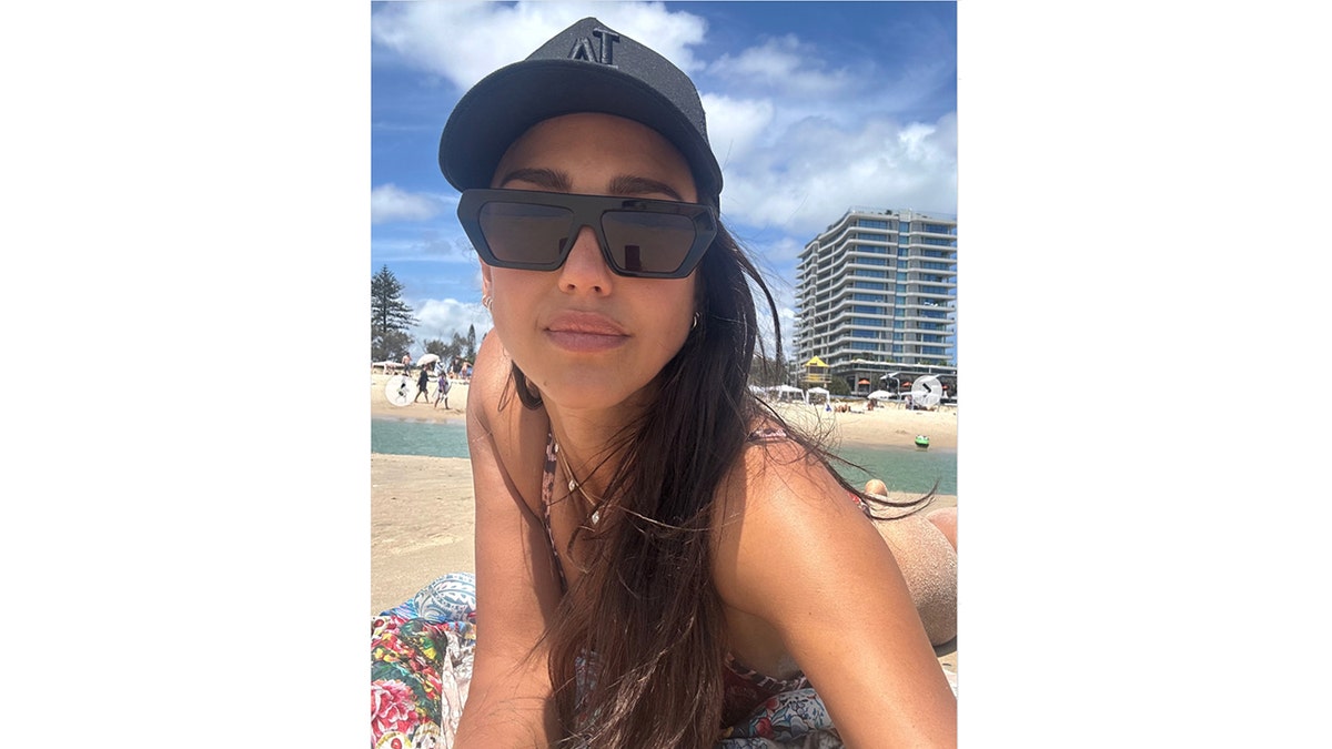 Jessica Alba takes a close-up beach selfie while lying on the sand in Australia, wearing oversized black sunglasses, a black baseball cap, and a patterned bikini top under a colorful coverup.